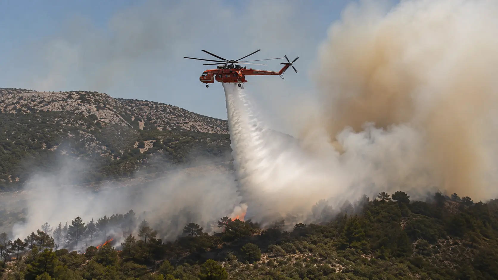 Firefighting helicopter making a water drop over a smoke-filled hillside during wildfire suppression operations, illustrating the low-altitude formation flying used in aerial firefighting