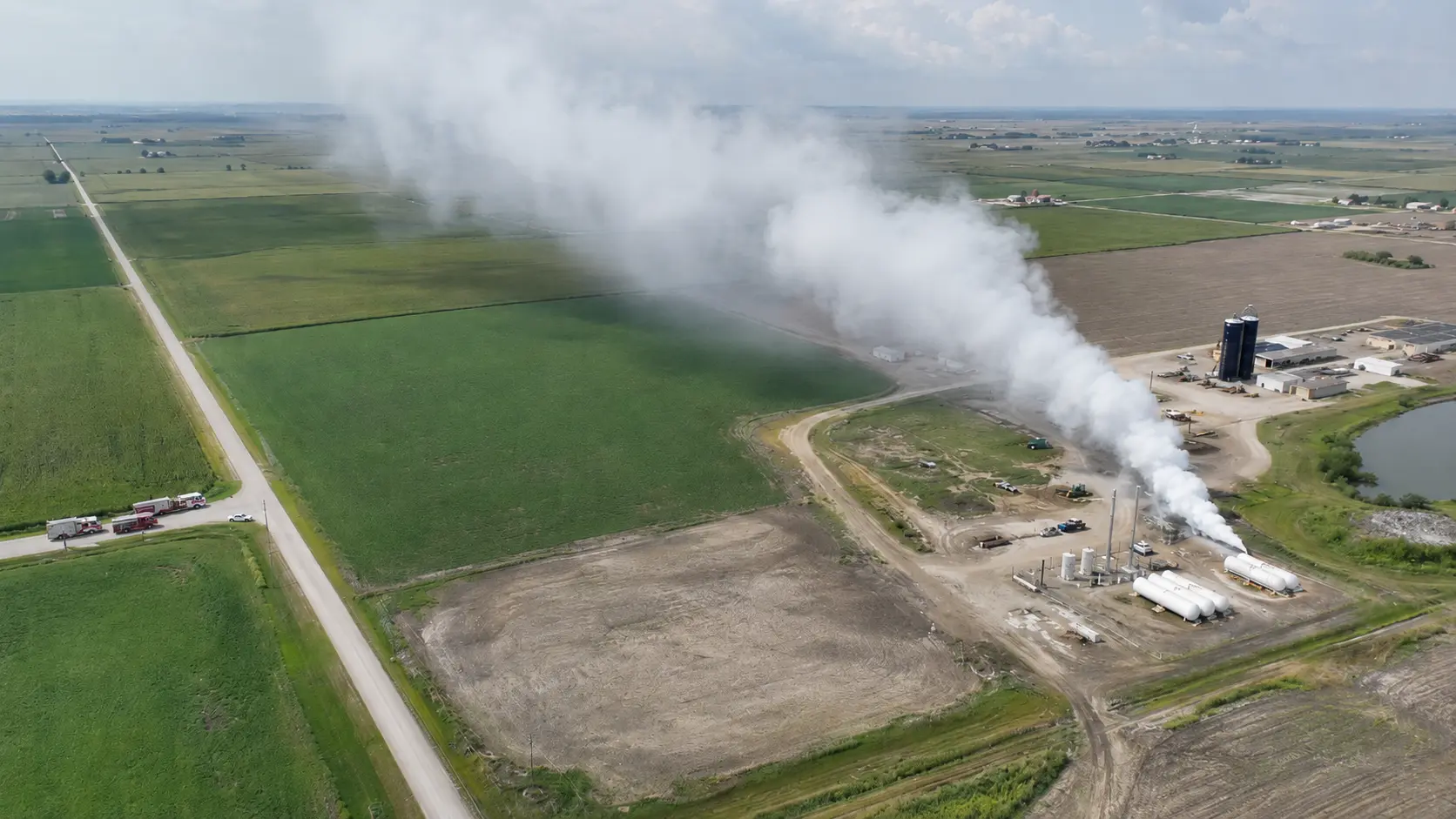 Photorealistic aerial photo showing an anhydrous ammonia release at an agricultural facility &mdash; a white vapor cloud visible rising from a storage tank area, clearly dispersing upward rather than settling to the ground (unlike heavier-than-air gases), fire department apparatus positioned upwind on a road approaching the facility, the cloud drifting downwind at elevated height &mdash; showing the lighter-than-air behavior of ammonia that rises and disperses differently from chlorine
