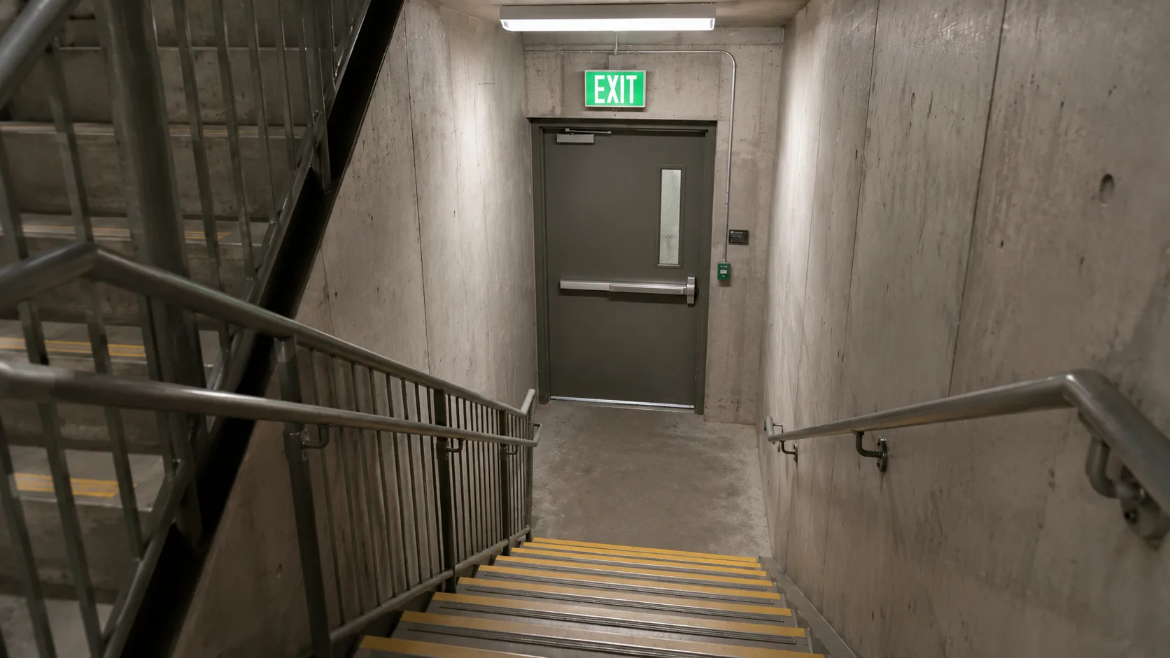 Illuminated green exit sign above fire stairwell door in apartment building corridor, showing clearly marked emergency stairwell with non-slip treads and handrails &mdash; the escape route renters must locate before a fire