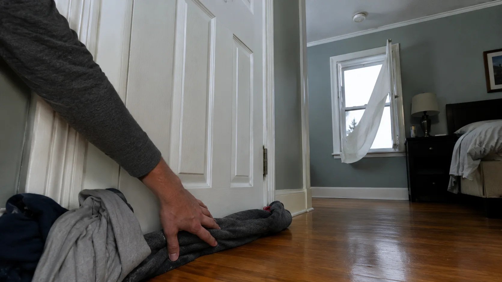 Person kneeling at a closed bedroom door pressing rolled towels and clothing firmly against the bottom door gap to seal against smoke infiltration during a house fire shelter-in-place situation, window visible in background slightly open &mdash; showing the correct fire survival technique when escape is blocked