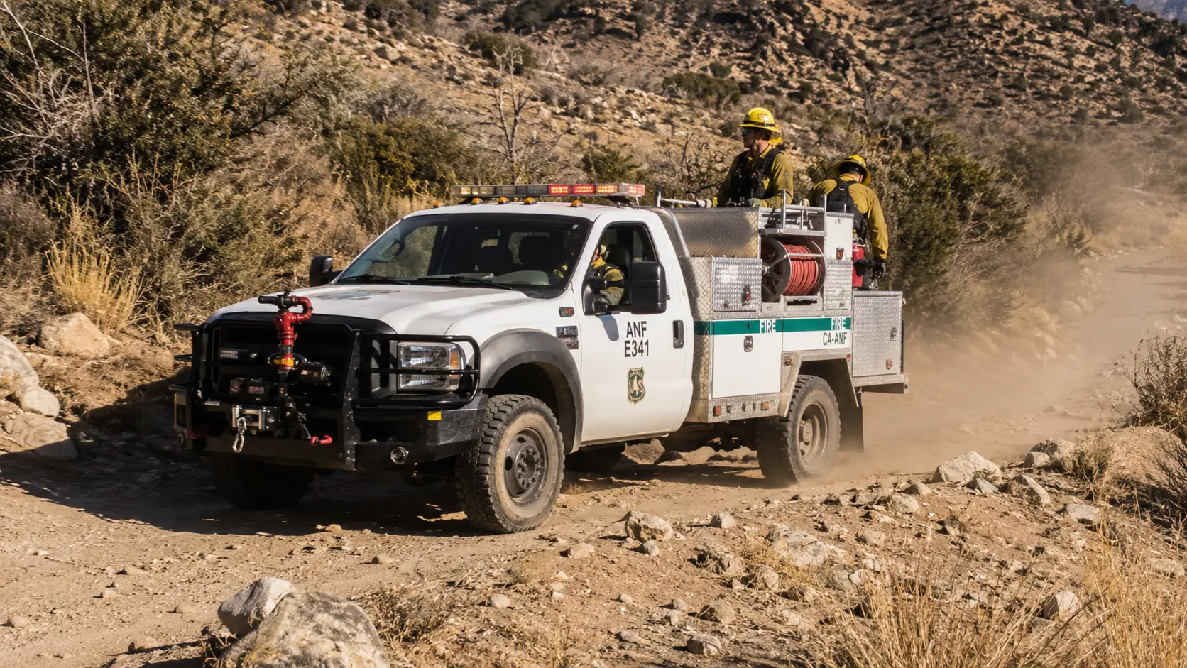 Photorealistic photo of a light brush truck (NIMS Type 6) &mdash; a pickup truck chassis with a small water tank body, approximately 150-gallon capacity visible through the tank window, a front-mounted bumper turret with a small nozzle, forestry hose reel on the side, and full all-wheel drive capability &mdash; positioned on a rocky unpaved off-road track through dry brush, showing the lighter more mobile configuration compared to a Type 3, two firefighters in Nomex visible
