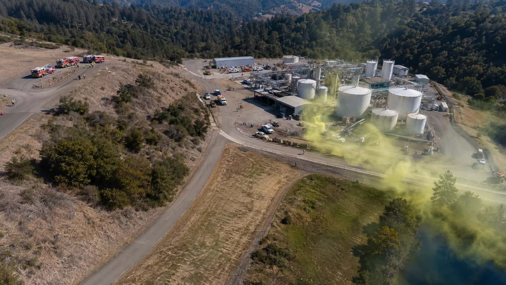 Photorealistic aerial photo showing a chlorine storage facility with a visible yellow-green gas plume drifting downwind and downhill from a release point, fire department apparatus positioned well upwind and uphill from the facility at the top of a slope, the gas plume clearly flowing away from the apparatus position &mdash; showing the correct upwind and uphill initial approach positioning for a chlorine gas release hazmat incident, industrial facility with storage tanks