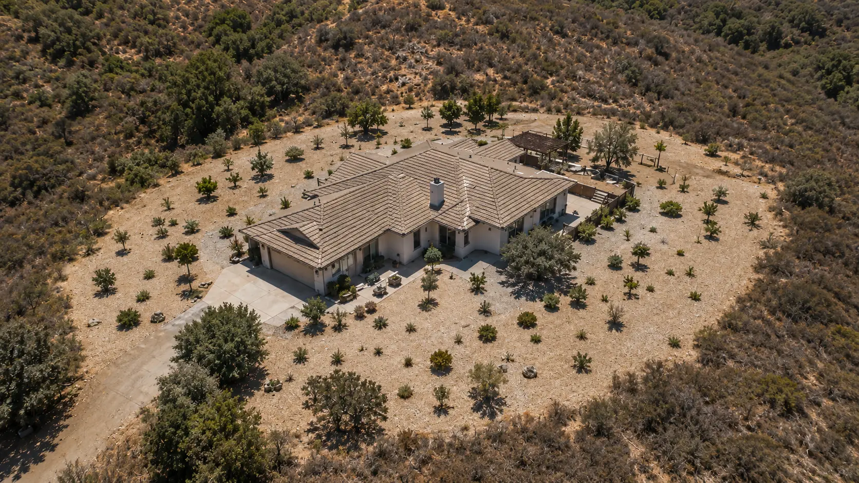 Photorealistic aerial drone photo of a well-maintained defensible space Zone 1 around a single-story home in a California foothill setting &mdash; showing the 30-foot cleared area around the structure with low irrigated plants spaced apart so their canopies do not touch, no continuous fuel path from ground to canopy or between plants, dry grass cleared from the immediate structure area, gravel mulch in Zone 0 visible at the foundation, the transition to less-managed natural brush visible at the 30-foot perimeter
