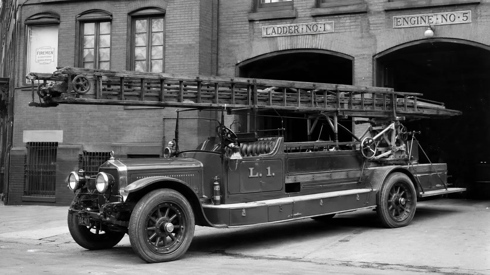 Early motorized ladder truck with a wooden extension ladder parked outside a fire station.