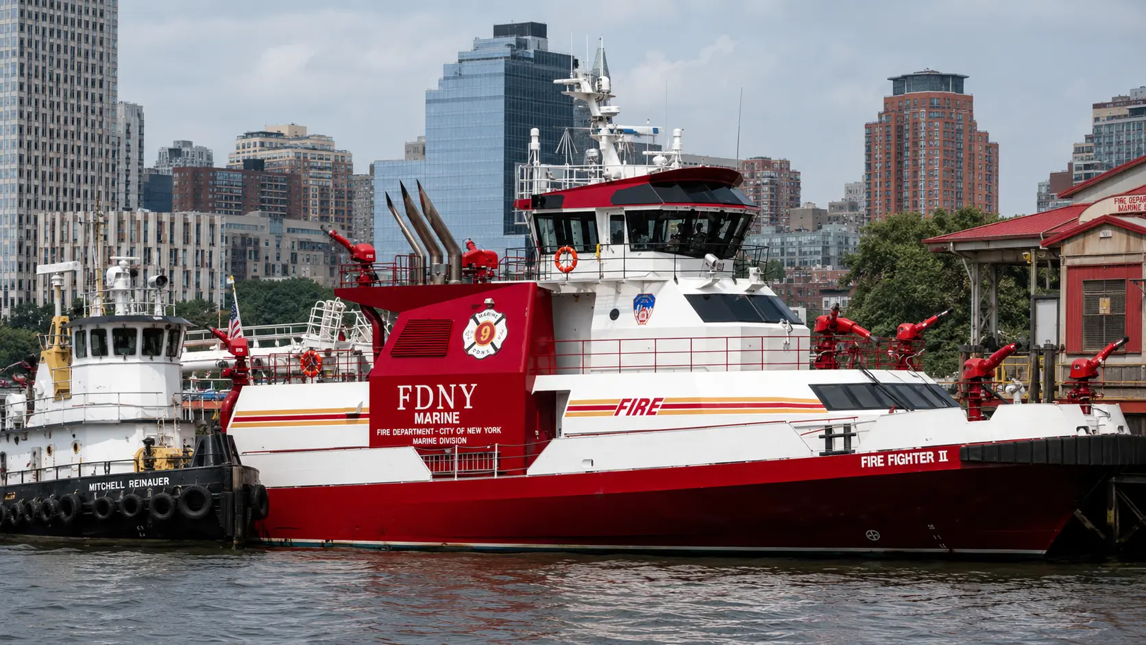 Photorealistic photo of FDNY's Fire Fighter II fireboat moored at its Manhattan pier station &mdash; a large red and white vessel approximately 130 feet in length, multiple monitor nozzles visible on the deck in raised position, the FDNY MARINE designation and Fire Fighter II name clearly visible on the hull, the Manhattan waterfront and skyline visible across the water in the background, the fireboat clearly visible as a substantial purpose-built marine fire apparatus rather than a converted vessel