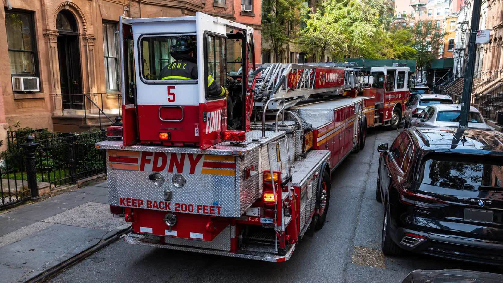 Photorealistic photo showing the tiller operator position at the rear of an FDNY tractor-drawn aerial ladder truck &mdash; a firefighter in FDNY turnout gear and helmet seated at the rear tiller steering wheel, using it to swing the rear of the long trailer around a parked car on a narrow New York City street to position the apparatus correctly in front of a fire building, the tractor cab visible ahead, urban NYC street scene