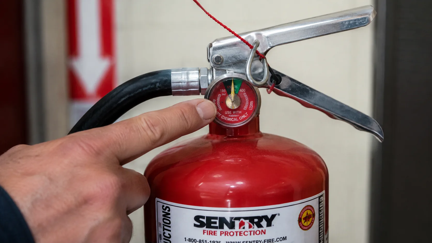 Close-up of fire extinguisher inspection showing a finger pointing to the pressure gauge needle in the green zone, the intact pull pin with tamper seal wire, and the condition label on the cylinder &mdash; the three key points of the monthly visual inspection that determines whether a fire extinguisher is ready for use