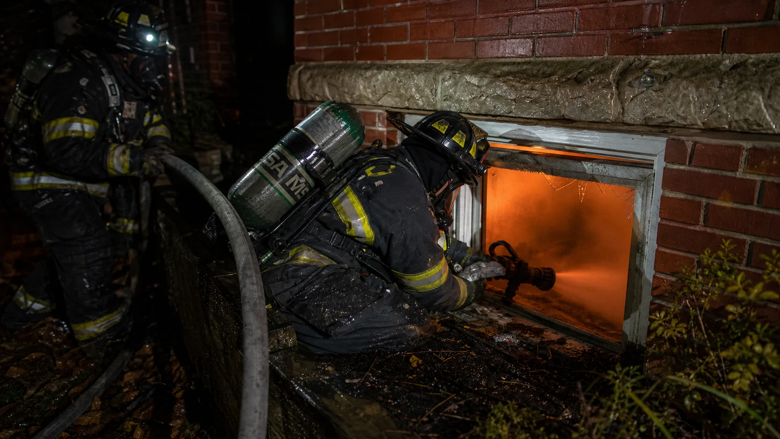 Photorealistic photo of a firefighter in full structural turnout gear and SCBA crouching at a below-grade basement window well, inserting a hoseline nozzle through the window opening to apply water to a basement fire &mdash; orange glow and smoke visible through the window, the firefighter in a low protected position at the window ledge, another firefighter behind holding the hoseline, realistic documentary fire service photography style, dramatic nighttime lighting