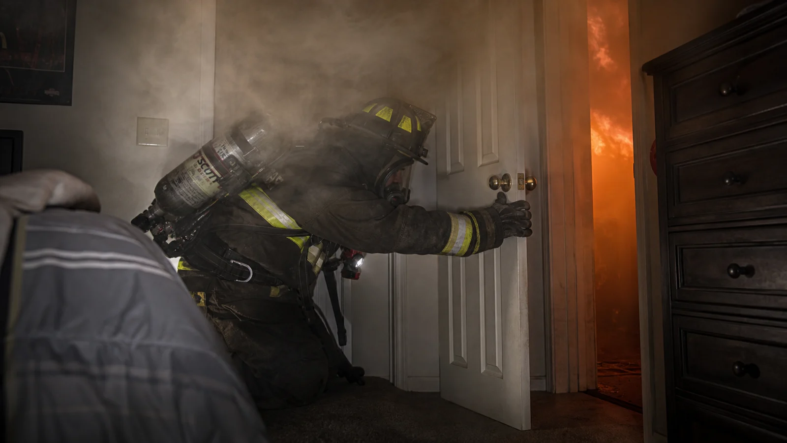 Photorealistic photo of a firefighter in full structural turnout gear and SCBA inside a smoke-filled bedroom reaching to close the interior door &mdash; the door partially open showing smoke and orange fire glow in the hallway beyond, the firefighter positioned low near the floor pushing the door shut &mdash; communicating the critical isolation step of the VEIS technique, realistic documentary fire service interior photography style, dramatic smoke and fire glow visible through the doorway gap