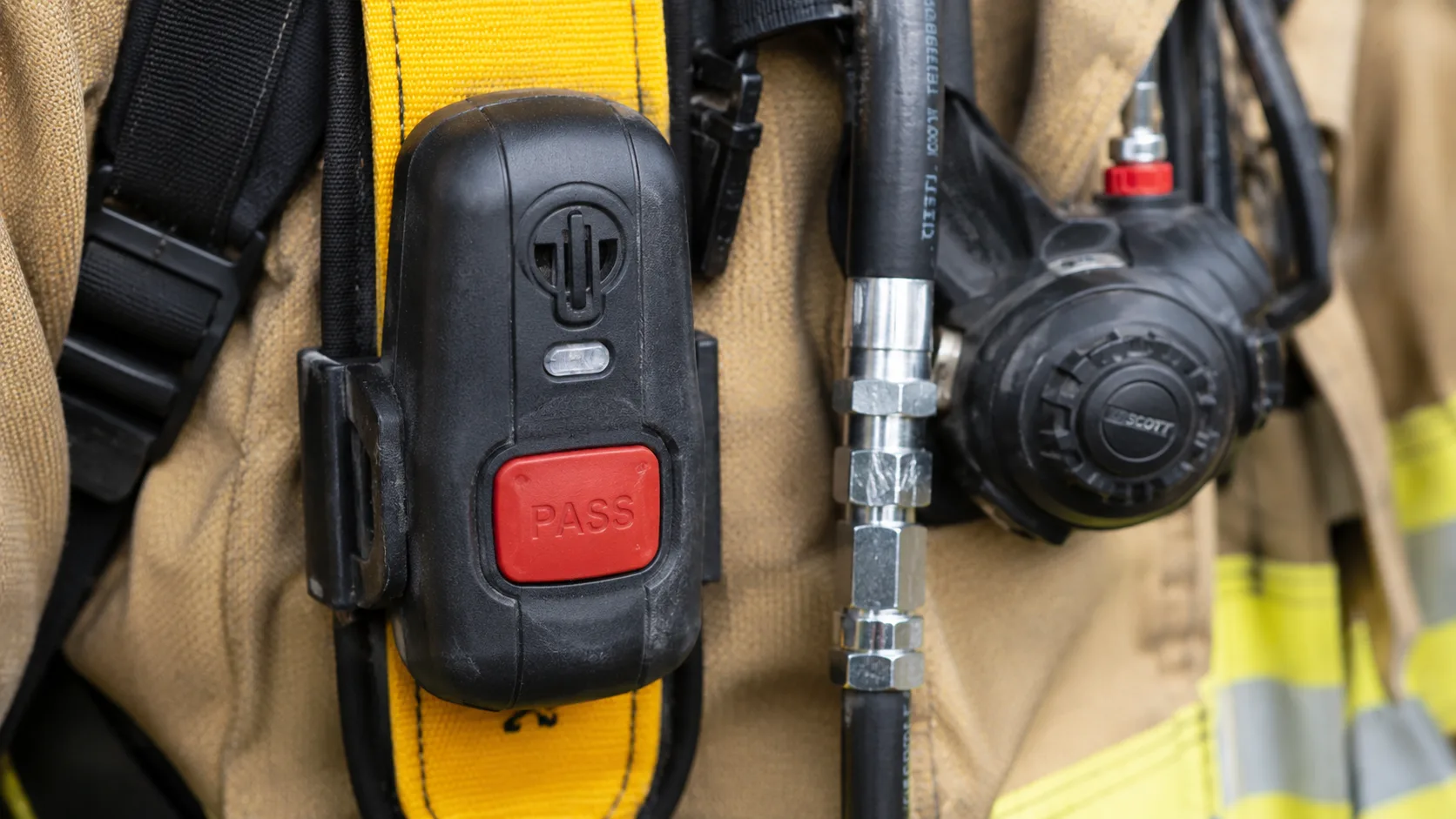 Close-up of a PASS (Personal Alert Safety System) device mounted on a firefighter's SCBA harness chest strap, showing the motion sensor housing and manual activation button, with SCBA cylinder and regulator visible in the background &mdash; the device that alerts rescuers when a firefighter has been motionless for 30 seconds