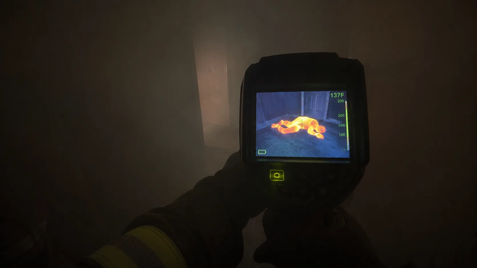 Point-of-view perspective of a firefighter holding a thermal imaging camera in a smoke-filled room, the camera screen showing a heat signature of a person lying on the floor in a bedroom doorway &mdash; demonstrating how thermal imaging cameras can detect victims in zero-visibility smoke conditions that are completely opaque to visible light