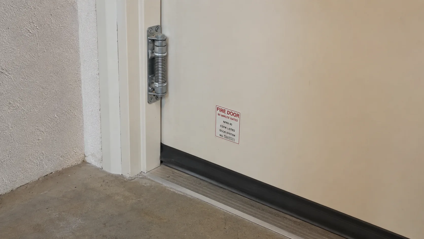Close-up of the interior side of a fire-rated door between an attached garage and house interior, showing the self-closing hinge mechanism, intact door sweep at the base, and fire door rating label &mdash; the critical barrier between garage fire and living space