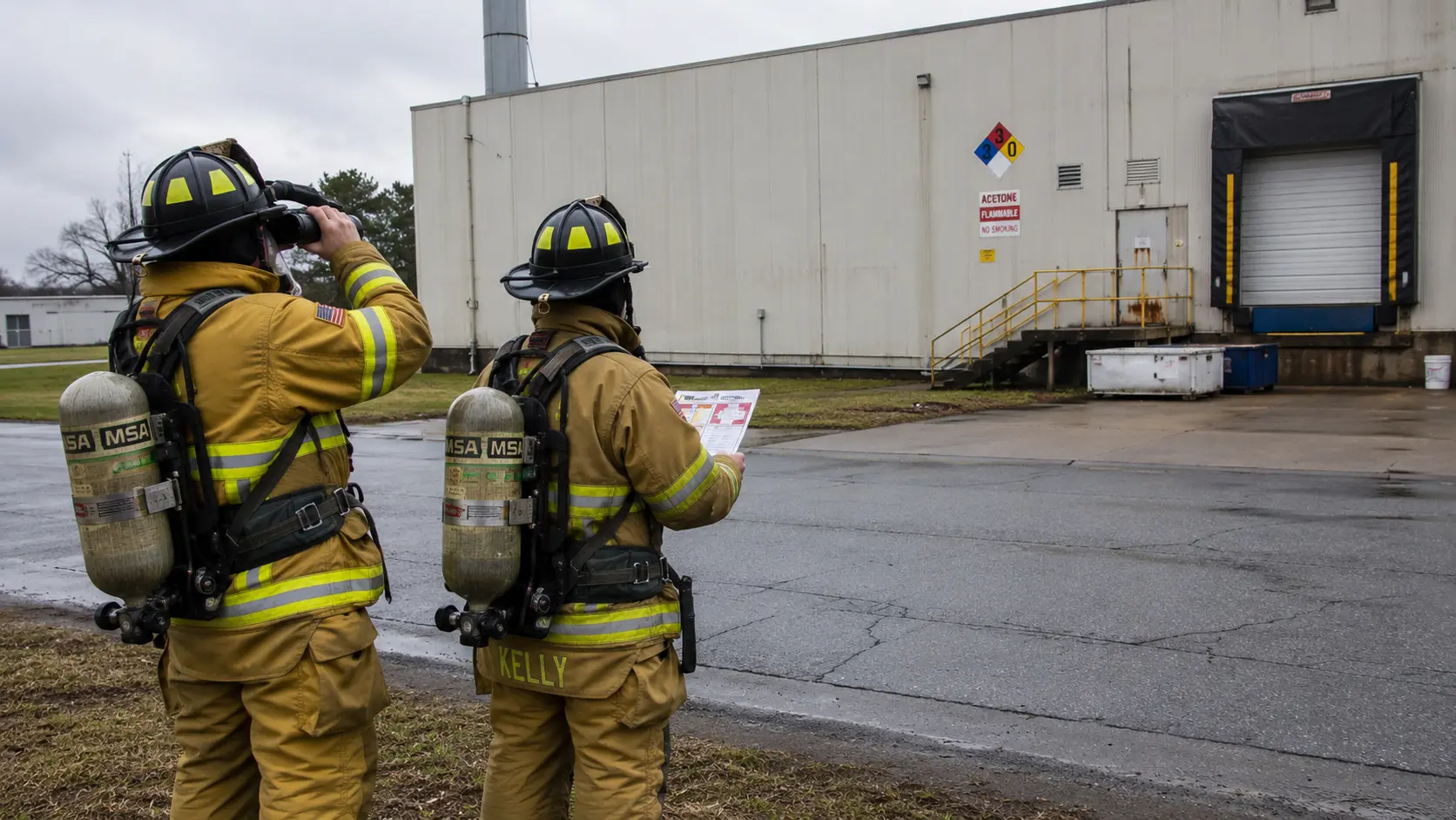 Photorealistic photo of two firefighters in full structural turnout gear and SCBA stopped at a safe distance from a chemical storage building, one firefighter holding binoculars and looking toward a large NFPA 704 diamond placard visible on the building wall approximately 100 feet away, the other firefighter consulting a hazmat reference guide &mdash; showing the approach assessment procedure before committing to operations at a hazardous materials incident