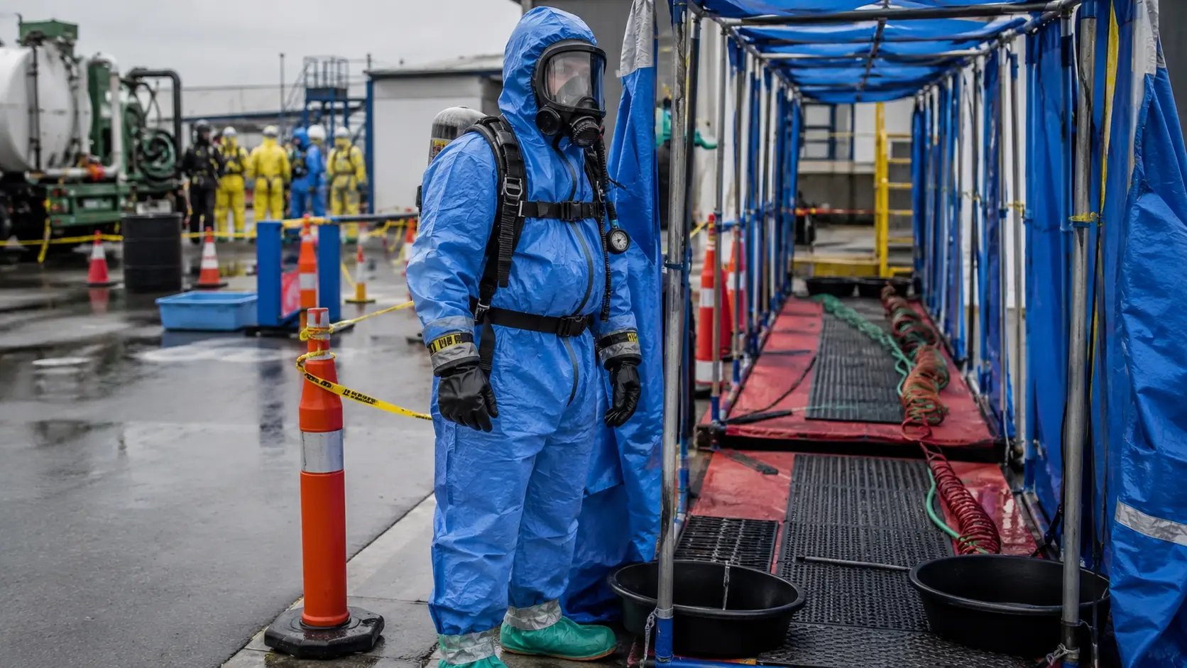 Photorealistic photo of a hazmat technician in Level B protection &mdash; a blue chemical splash-protective coverall suit with taped seams at the wrists and ankles, SCBA worn on the outside of the suit with the harness over the chemical suit, full-face SCBA mask, chemical-resistant outer gloves, wearing protective boot covers &mdash; standing at the edge of a marked warm zone at an industrial chemical spill scene, decontamination corridor visible in the background