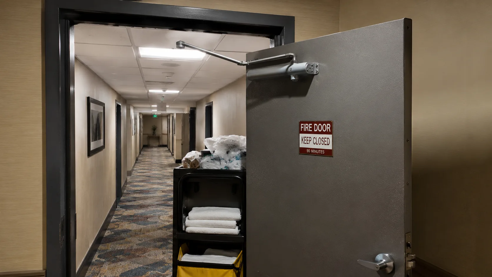 Hotel corridor fire door propped open with a luggage cart, showing the self-closing mechanism on the door frame and the fire door rating label &mdash; illustrating the fire safety hazard of keeping corridor fire doors from closing in a hotel building