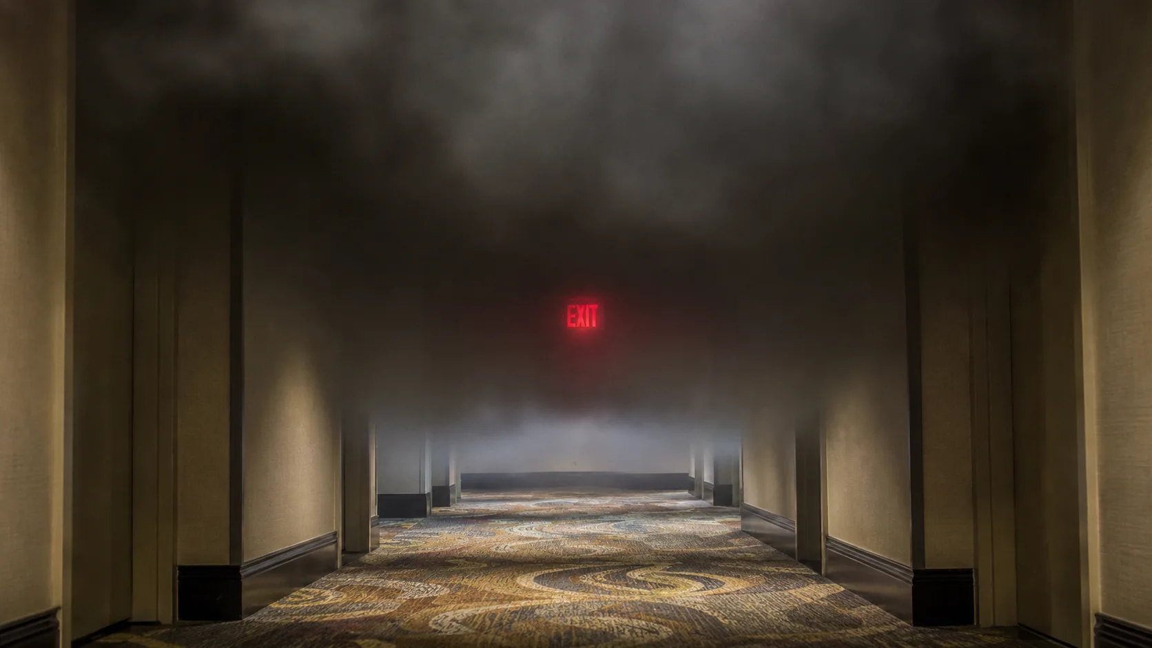 Hotel corridor filling with dark smoke near the ceiling, showing the clear air layer at floor level below the smoke line &mdash; demonstrating the low-crawl survival technique where guests should stay below the smoke while evacuating through a hotel hallway fire emergency