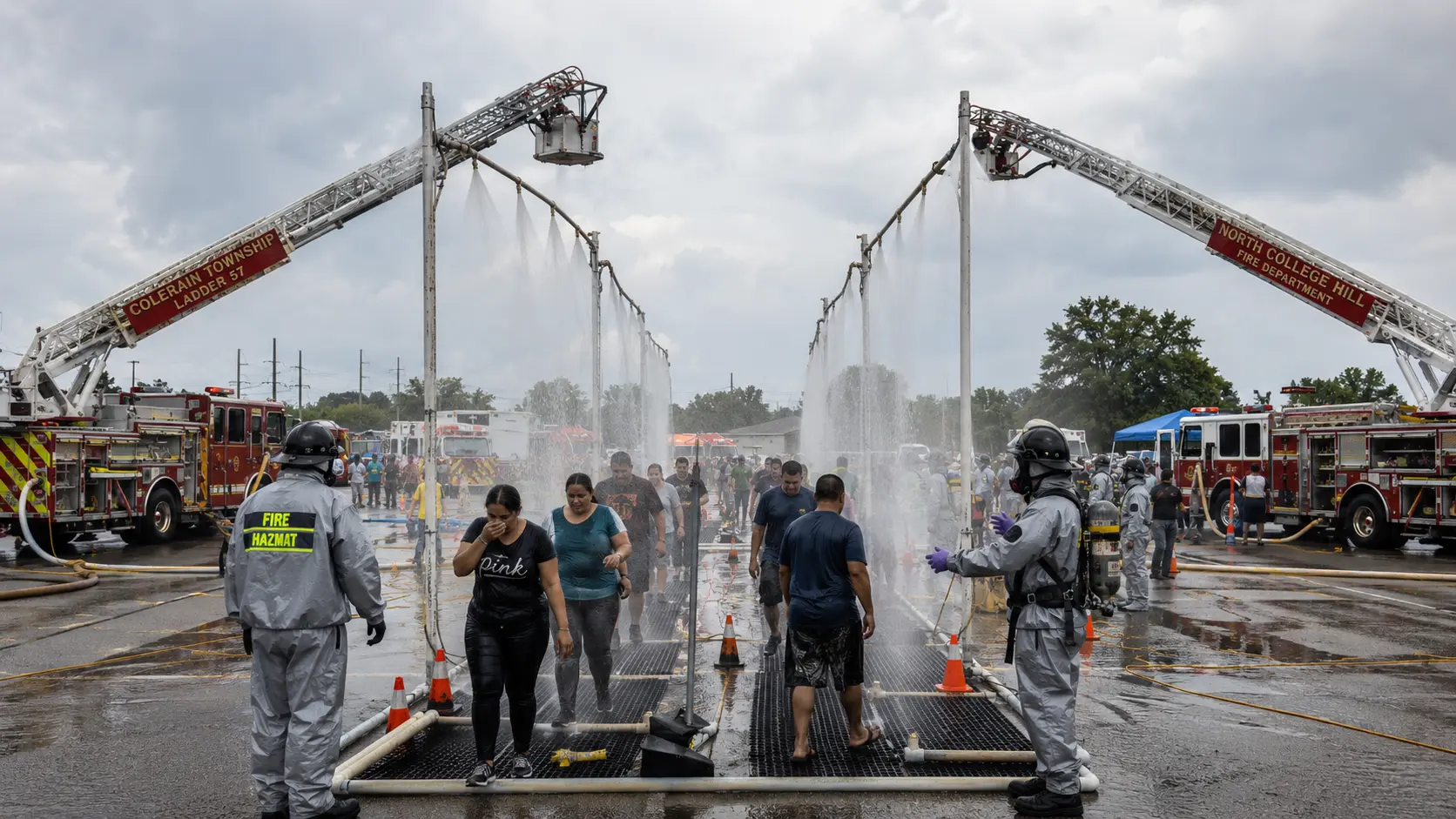 Photorealistic photo of a mass decontamination operation for civilians &mdash; a long corridor created by two fire engines facing each other with ladder-mounted water spray systems creating parallel water curtain 'showers,' multiple civilians in street clothes walking through the water corridor to rinse off contamination, fire department personnel in Level C protection directing people through the line, a large-scale outdoor setup suggesting a chemical release mass casualty scenario, 16:9 ratio