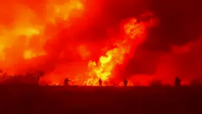Firefighters in protective gear battling intense wildfire with hoses amid smoky, burning forest landscape.
