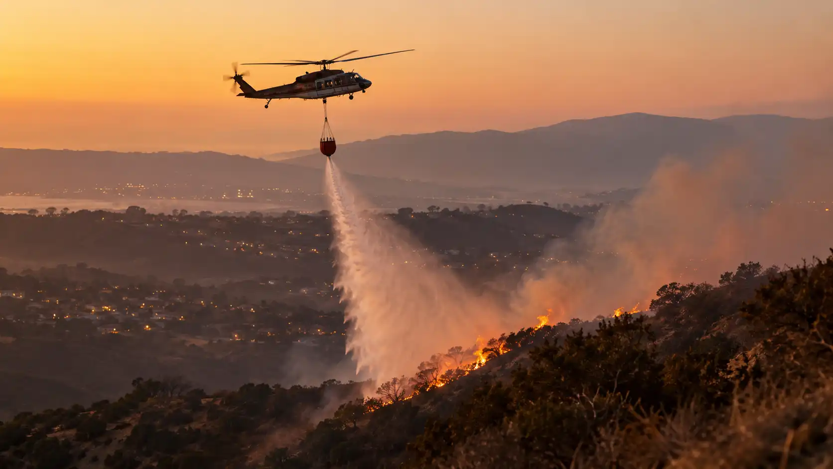 A fire helicopter dropping water on a small brush fire on a hillside at dawn