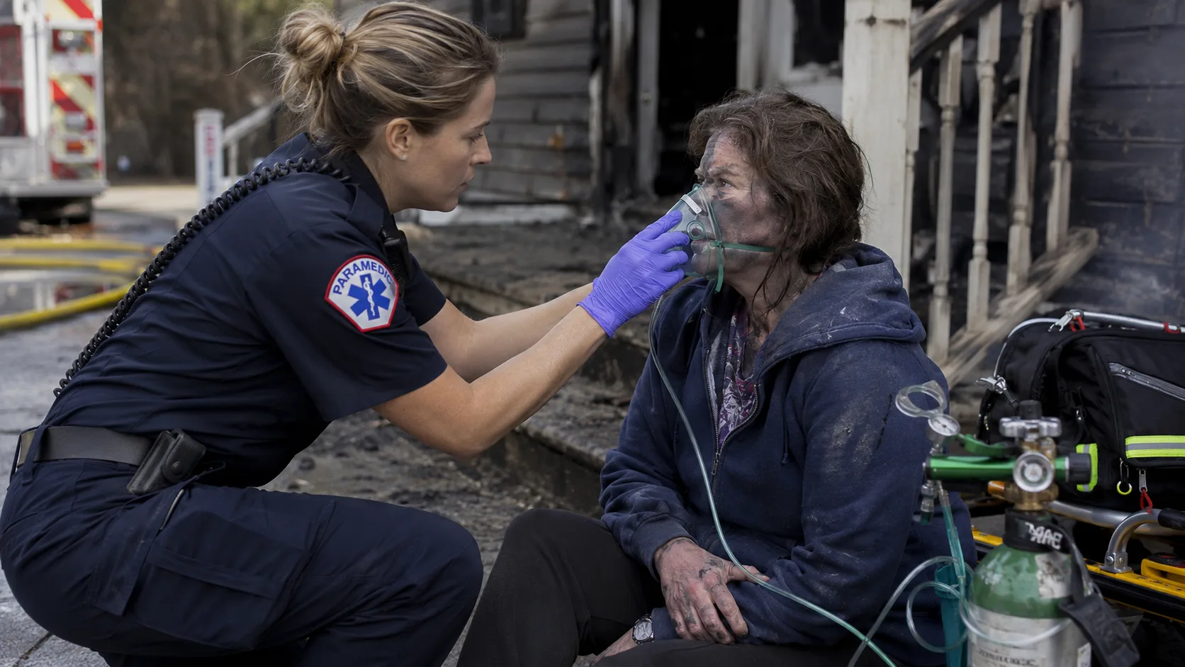 Paramedic placing a non-rebreather oxygen mask on a fire survivor sitting at the exterior of a house fire scene, the person showing mild soot on face and clothing, EMS crew visible in background &mdash; high-flow oxygen administration at the scene is the first treatment for suspected carbon monoxide poisoning from smoke inhalation