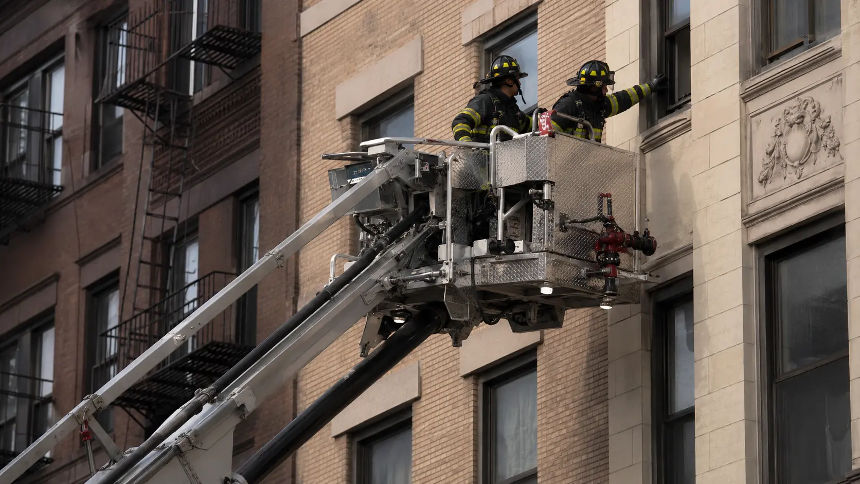 Photorealistic photo of a fire department tower ladder with its aerial elevated and the large platform basket positioned at the fourth floor window of a residential apartment building, two firefighters in full turnout gear visible working in the basket &mdash; one operating the basket controls while the other reaches toward the window opening, the hydraulic boom and turntable visible below, realistic basket guard rails and monitor nozzle mounted at the basket front, urban setting