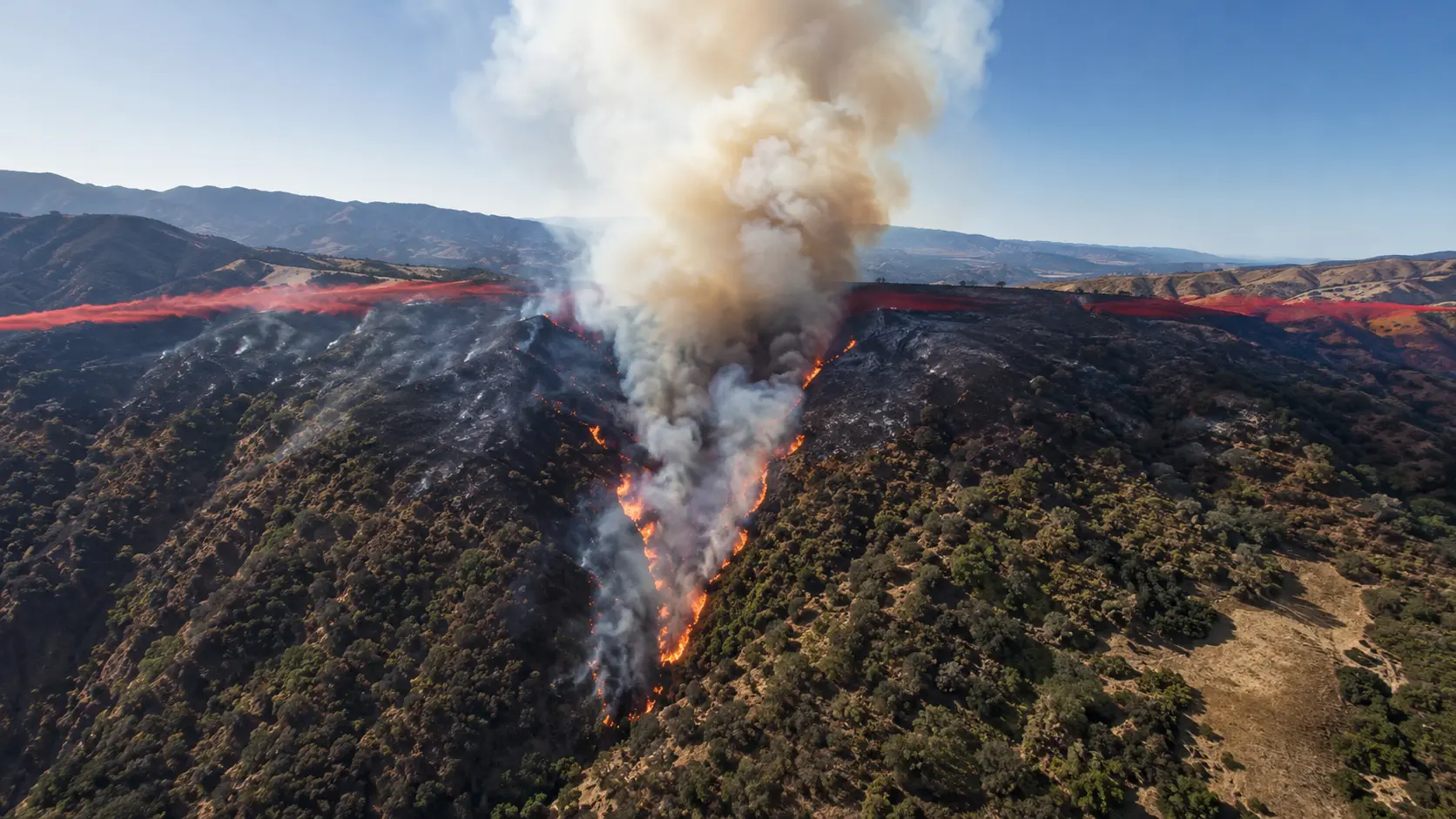 Aerial photograph taken from a helicopter showing a fast-moving wildfire burning up a steep chaparral-covered canyon slope in Southern California &mdash; the fire front clearly visible as an advancing orange line moving uphill with wind-driven flames leaning forward in the direction of spread, white smoke column rising from the head of the fire, burned black area visible below, unburned tan-colored dry brush ahead of the fire front, mountain ridgeline visible in the background under a hazy smoke-filled sky