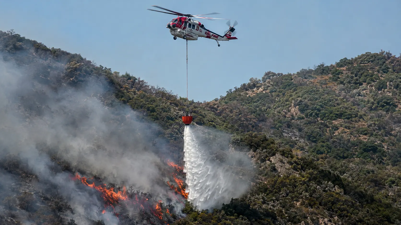 Helicopter releasing water from a suspended bucket over a hillside wildfire.